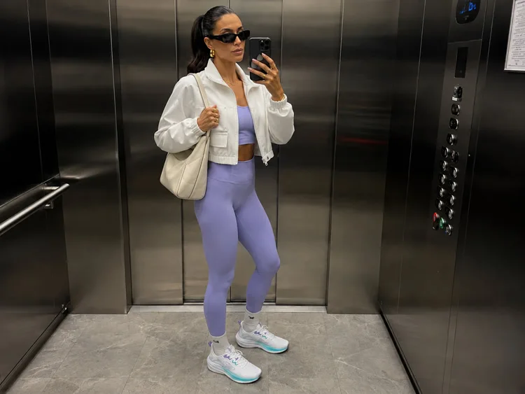 Mulher usando look athleisure pós-treino com legging lilás, top esportivo, jaqueta branca, tênis feminino da Pegada e bolsa em espelho de elevador.