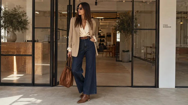 mulher aparece em um ambiente de trabalho moderno, com uma proposta de look elegante e contemporânea. Ela usa blazer bege, blusa clara com caimento fluido, jeans de modelagem ampla, cinto em tom terroso e bolsa marrom, compondo uma produção equilibrada entre sofisticação e casualidade. Nos pés, o destaque fica para a bota marrom de cano curto da coleção de inverno da Pegada, que complementa o visual com personalidade, conforto e um toque refinado, ideal para inspirações de looks para trabalhar.