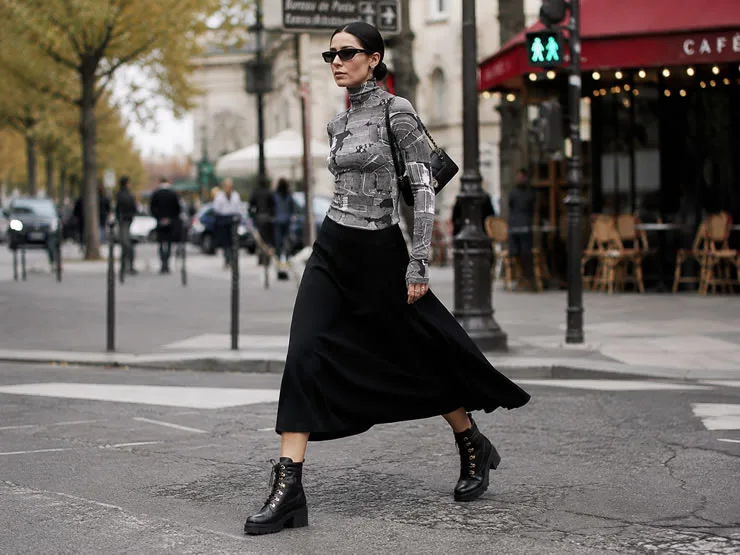 Mujer caminando por una calle urbana usando blusa de cuello alto estampada en tonos de gris, falda midi negra y borcegos negros de Pegada.