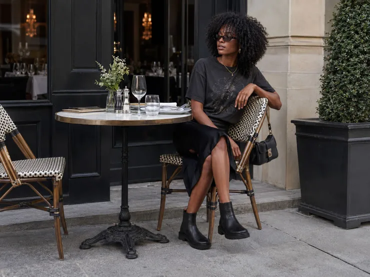 Mulher preta com cabelo crespo sentada em café elegante, usando camiseta gráfica escura, saia preta, bolsa pequena e bota preta da Pegada.